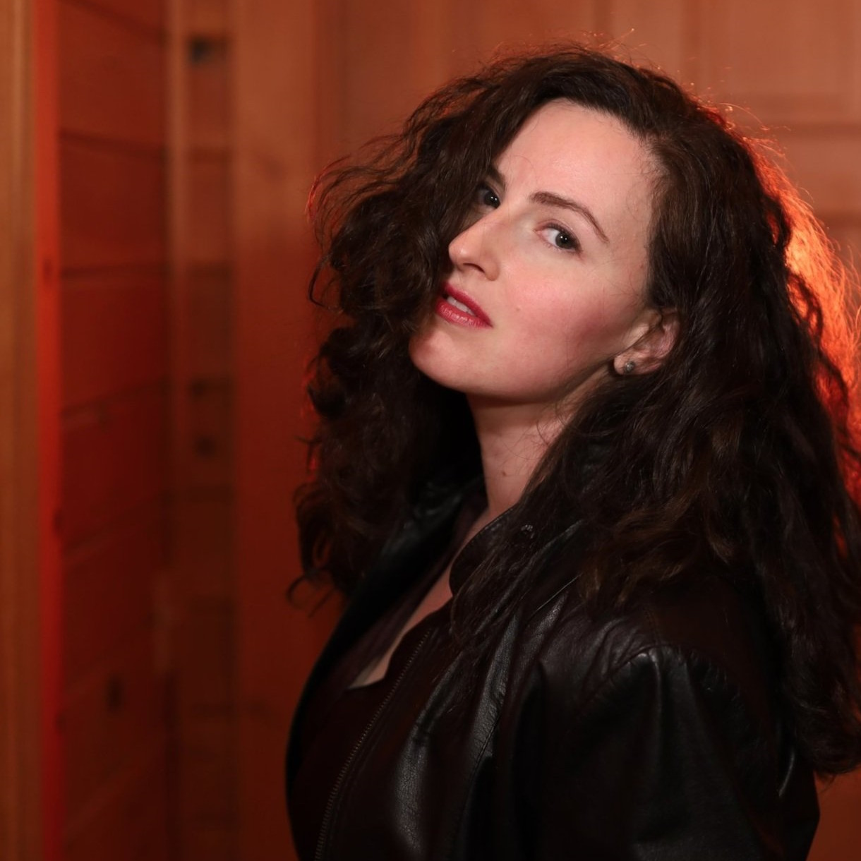 A young woman with long, dark, wavy hair faces the camera over her shoulder. She is wearing a dark leather jacket.